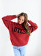 A female model wearing a red turtleneck sweater with 'Utes' text on the front, and a smile on her face.