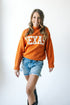 A woman wearing an orange turtleneck sweater with the text 'TEXAS' across the front, paired with denim shorts.