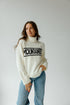 Woman wearing a white sweater with 'Mountaineer' text and blue jeans on a plain background