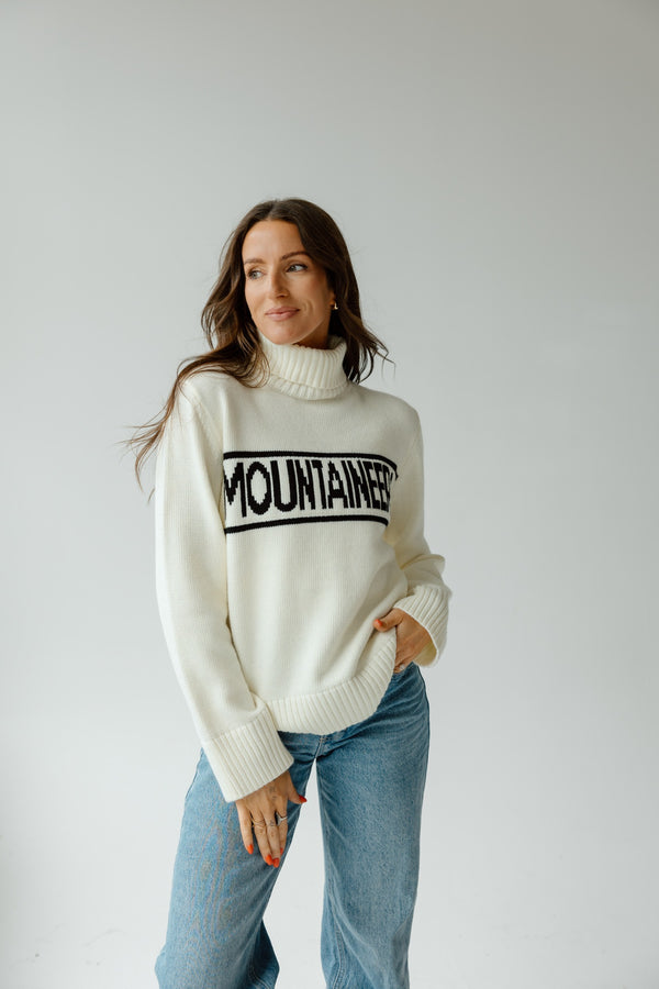 Woman wearing a white sweater with 'Mountaineer' text and blue jeans on a plain background