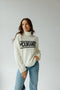 Woman wearing a white sweater with 'Mountaineer' text and blue jeans on a plain background