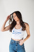 Woman wearing a white tank top with blue text and blue jeans against a white background